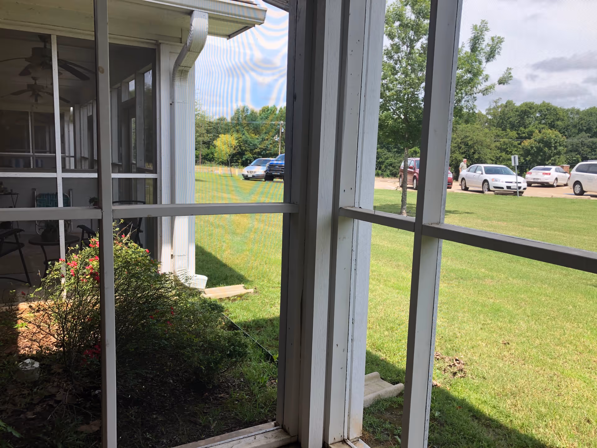 View through a screened porch looking out onto a grassy lawn, parked cars, and trees.