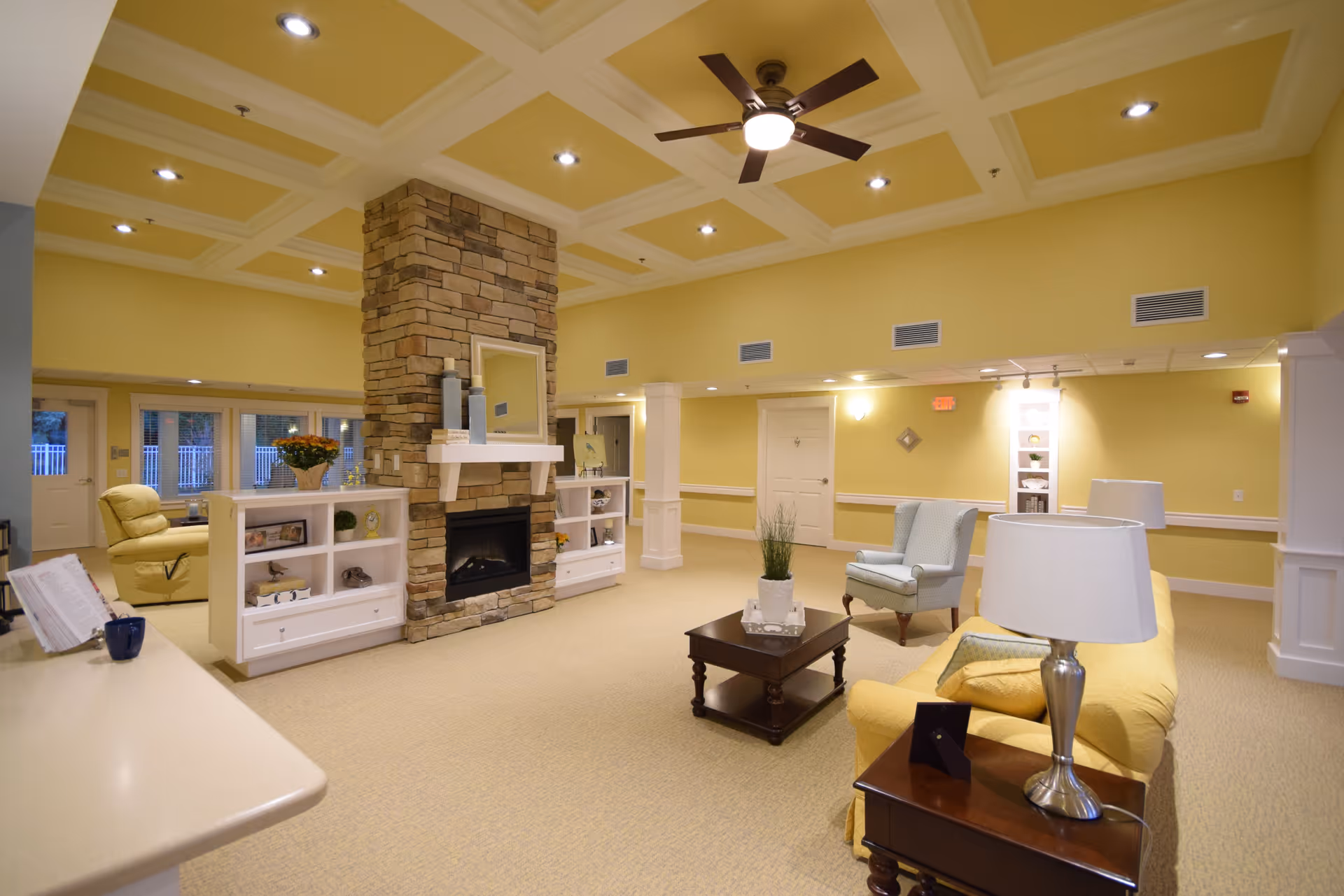 A spacious senior living common area with yellow walls and beige carpet. The room features a stone fireplace with a white mantel, flanked by built-in white shelves. There is a yellow sofa with a side table and lamp, a blue armchair, and a dark wooden coffee table with a plant on it. The ceiling has recessed lighting and a ceiling fan with a light. Large windows and doors are visible in the background.