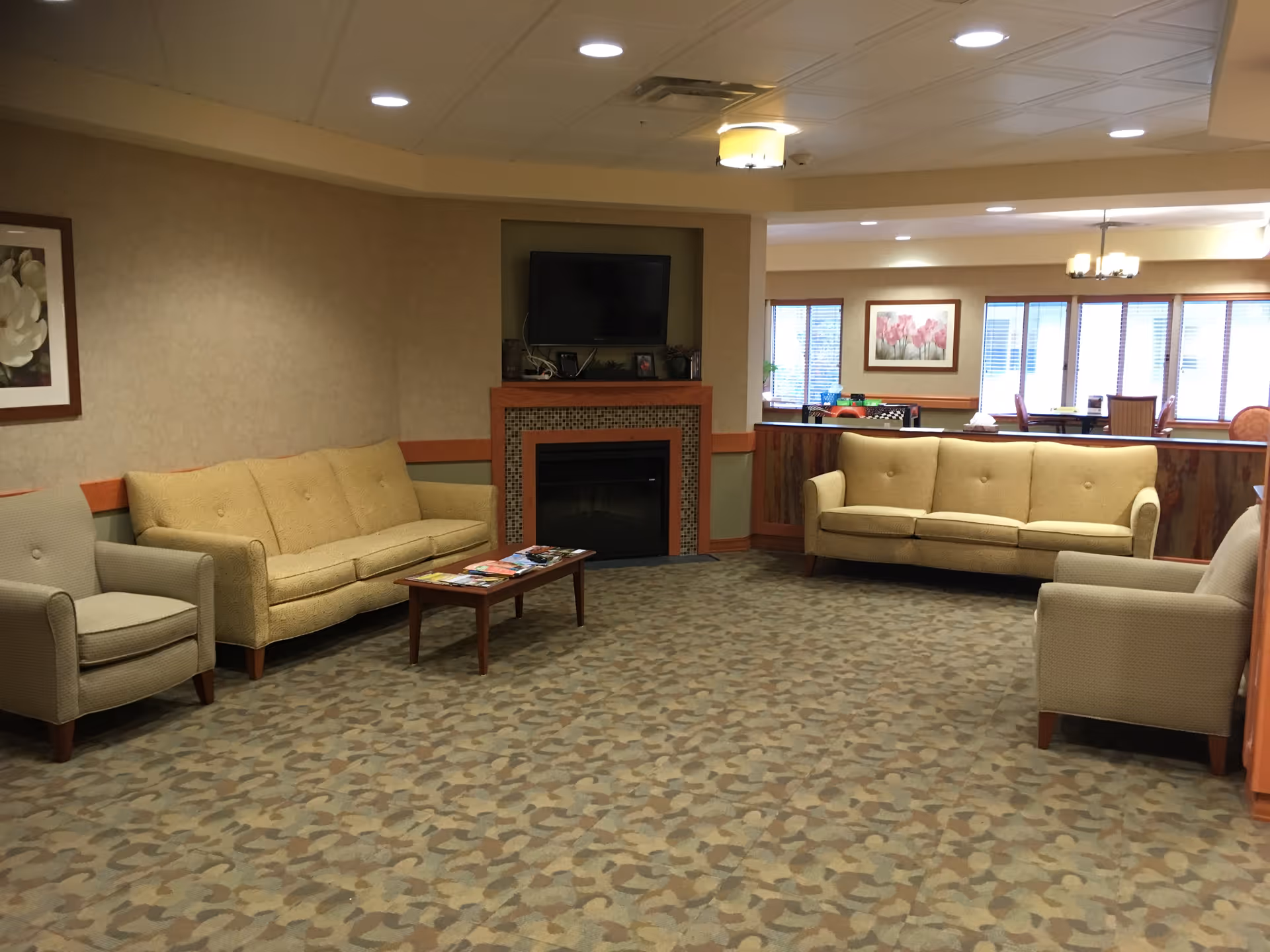 A comfortable common lounge with sofas and armchairs around a coffee table, a fireplace with a mounted TV, and patterned carpet.
