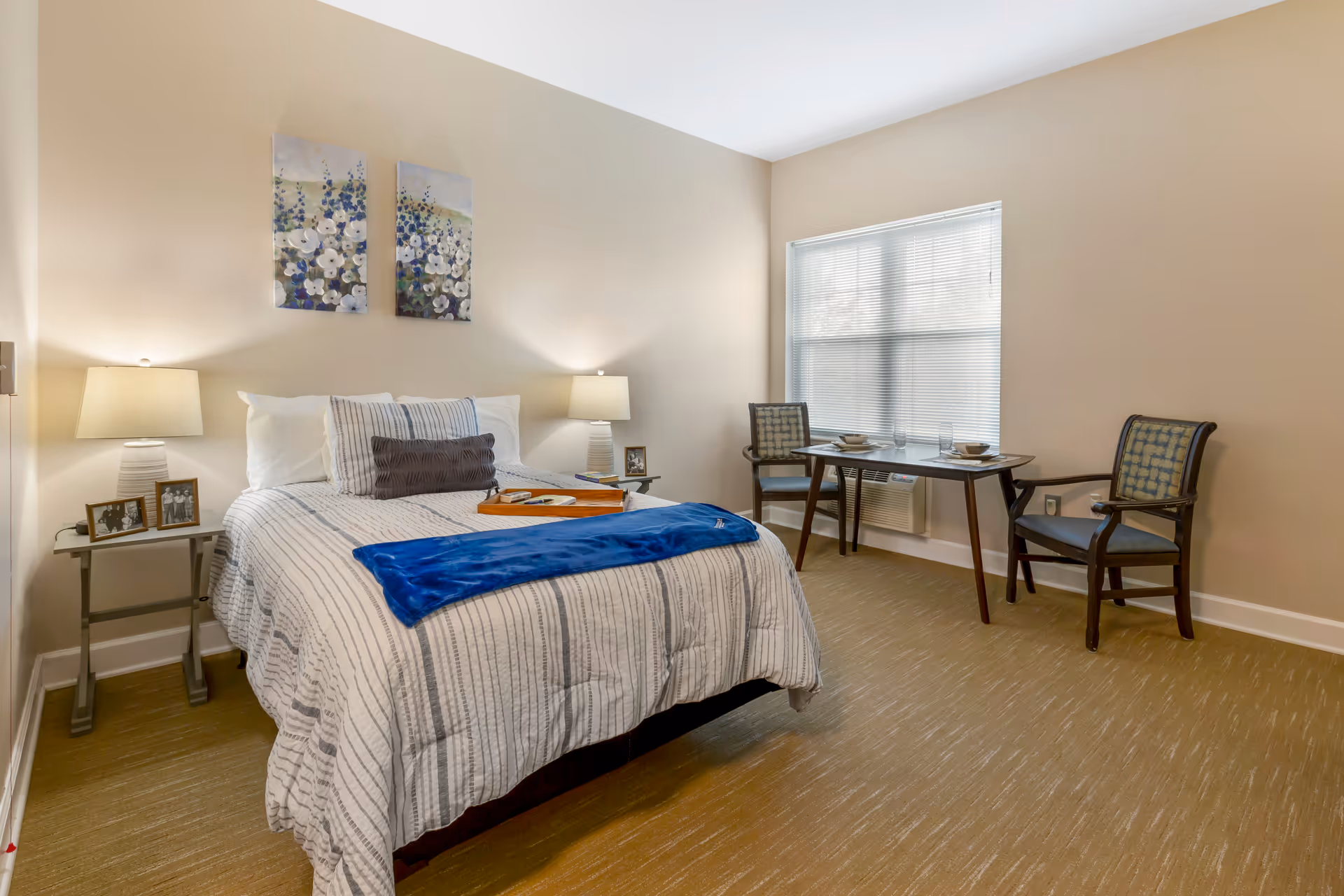 Neatly furnished bedroom with a made bed, bedside tables and lamps, artwork above the bed, and a table with two chairs by a window.