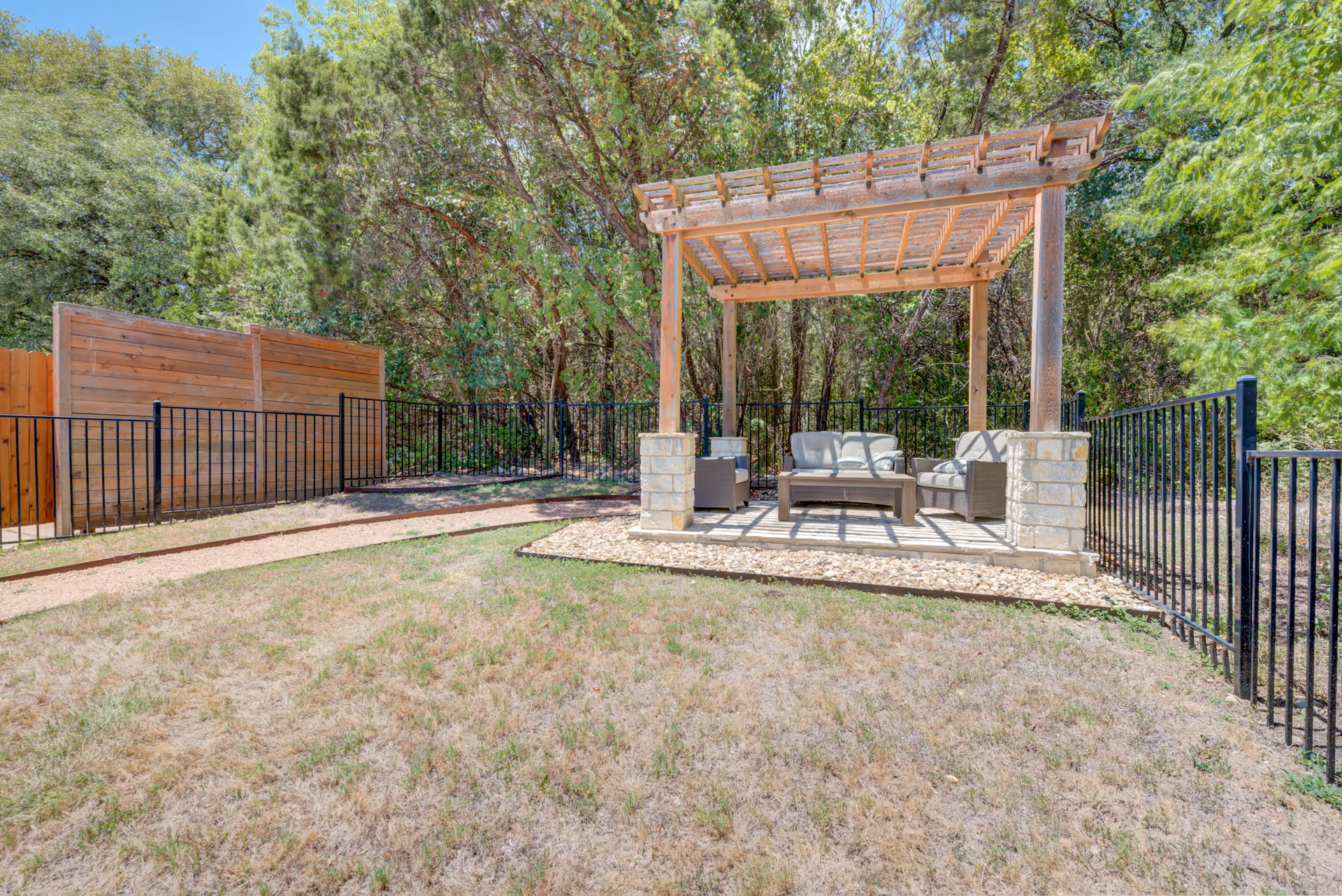 Outdoor seating area with a wooden pergola covering cushioned chairs and a coffee table, surrounded by a black metal fence and trees in the background under a clear blue sky.
