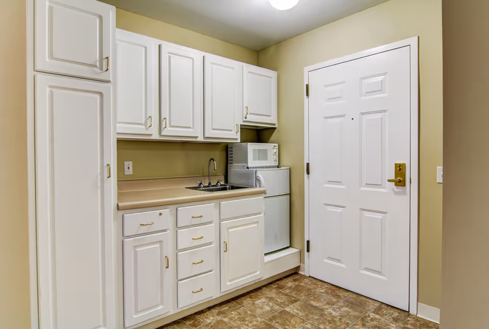Small kitchen area with white cabinets, a countertop with a sink, a microwave on top of a mini refrigerator, and a white door to the right. The floor is tiled and the walls are painted beige.