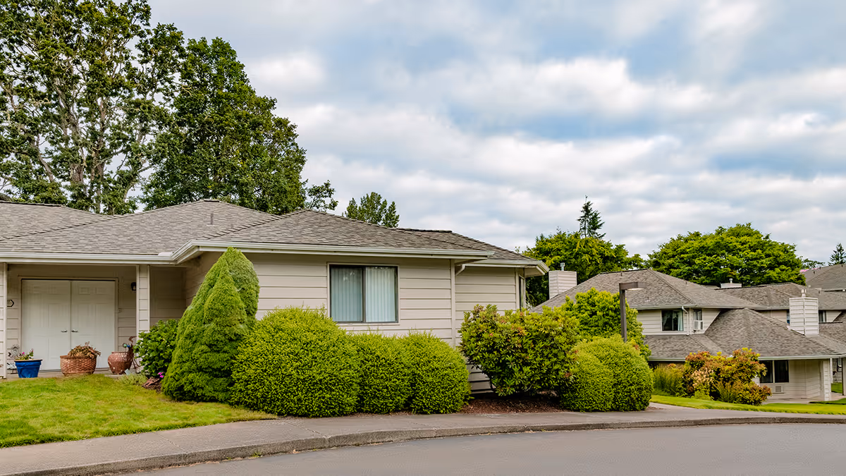 Single-story beige residential building with shrubs and a manicured lawn under a cloudy sky.