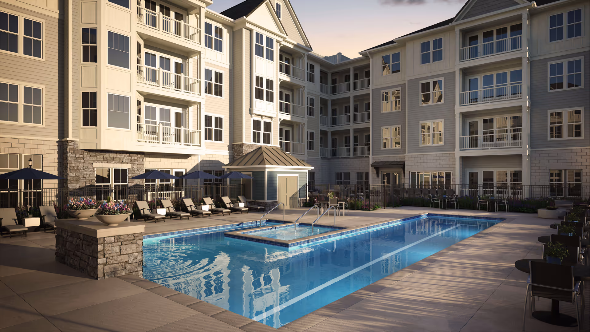 Outdoor swimming pool area at a residential facility with lounge chairs, umbrellas, tables, and chairs surrounding the pool. The pool is rectangular with a smaller square section in the middle. The building in the background has multiple floors with balconies and large windows.