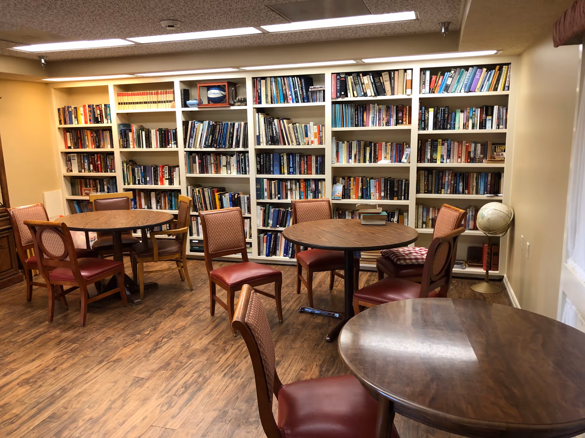 A cozy library room with wooden floors, several round wooden tables surrounded by cushioned chairs, and white bookshelves filled with books along the back wall. A globe stands on a stand in the corner near the window with sheer curtains.