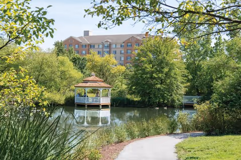 A gazebo on a pond with a walking path and trees in the foreground and a multi-story senior living building in the background.