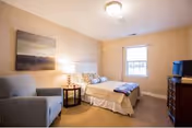 A cozy bedroom with a bed featuring white bedding and decorative pillows, a gray armchair, a round wooden side table with a lamp, a window with blinds, a wooden dresser with a small TV on top, and a landscape painting on the wall.
