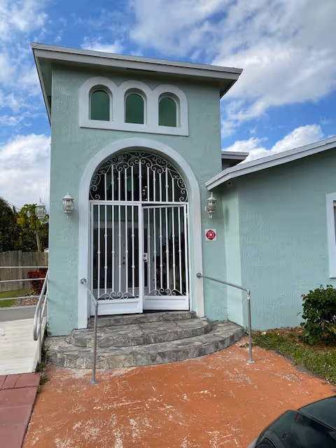 Entrance of a light green building with a decorative white metal gate and arched windows above. There are stone steps leading up to the door with metal handrails on both sides. The sky is partly cloudy.