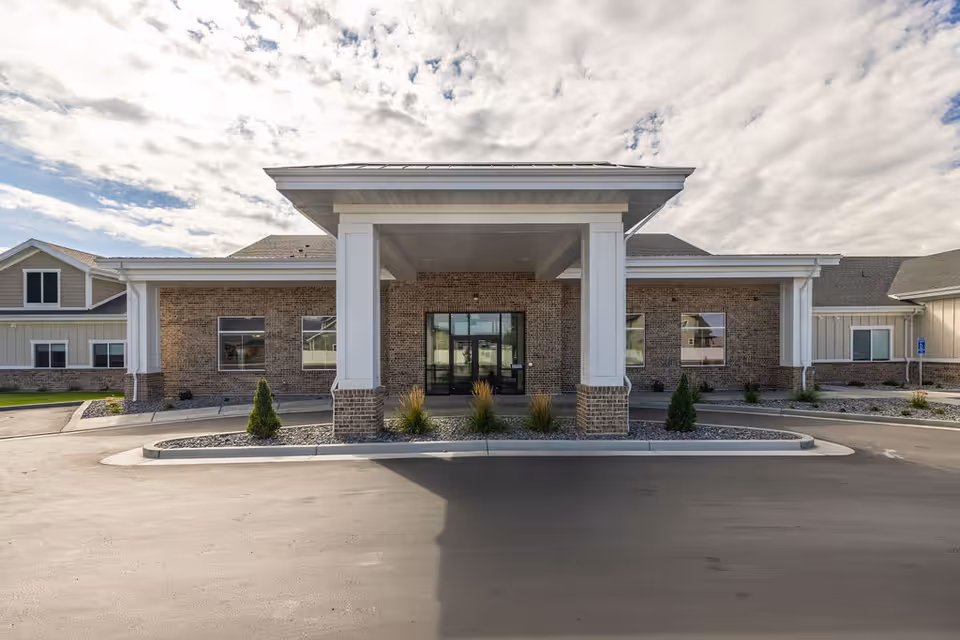 Front exterior view of a brick assisted living facility with a covered entrance and landscaped driveway.