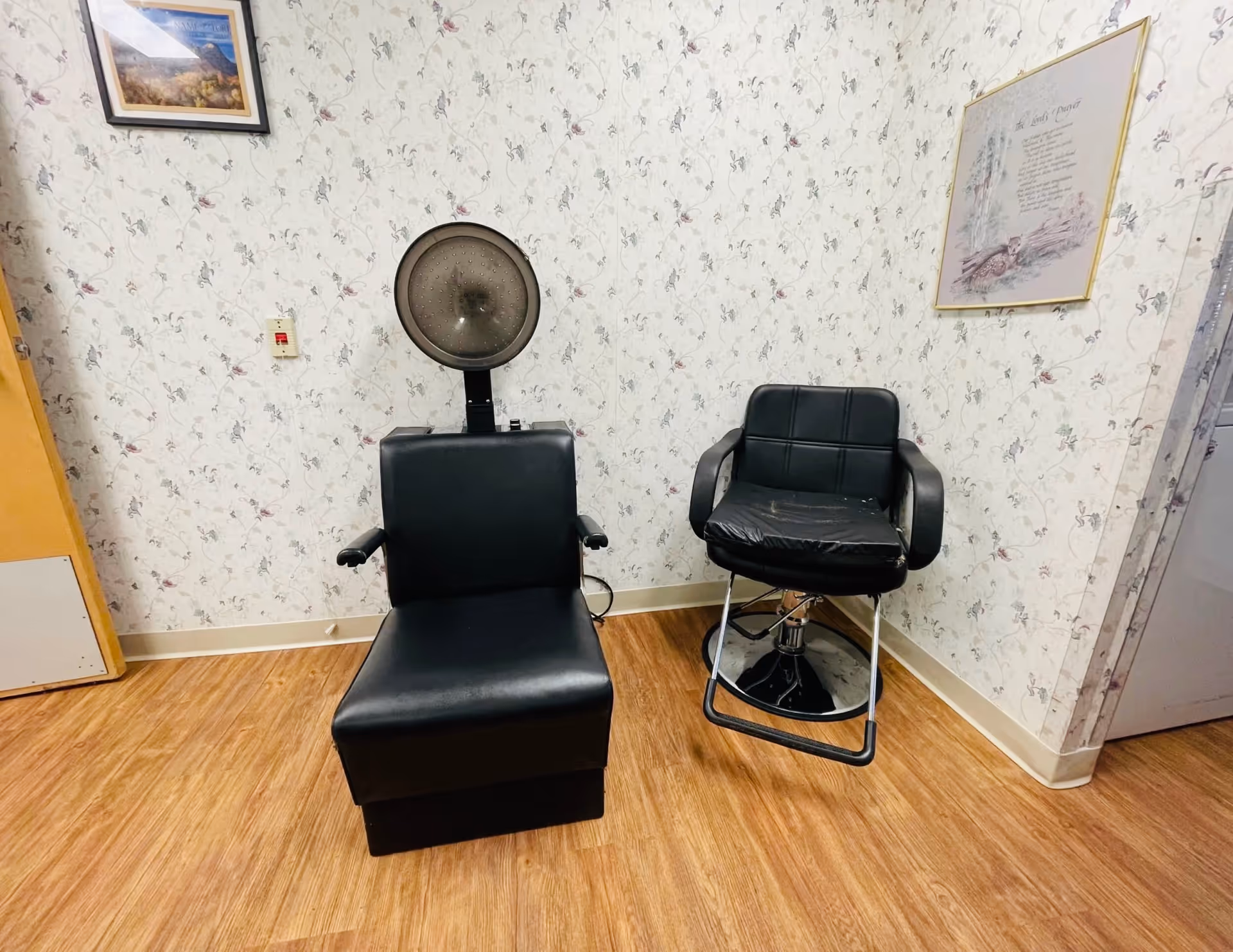 Two black salon chairs, one with a hooded hair dryer, against floral wallpaper in a small room with wood flooring.