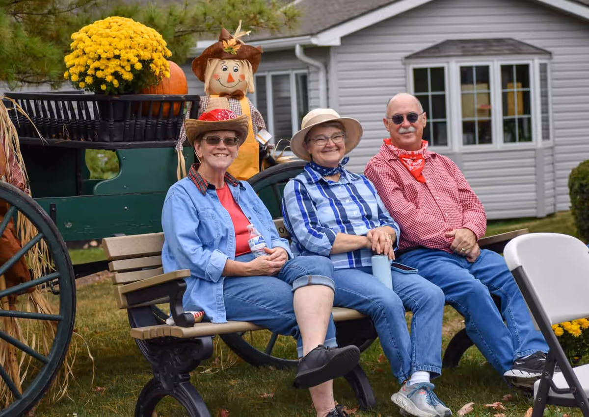 Three elderly people sitting on a bench outdoors in front of a building. They are dressed casually with hats and bandanas, smiling and enjoying the day. Behind them is a decorative wagon with a scarecrow, a pumpkin, and a pot of yellow flowers.