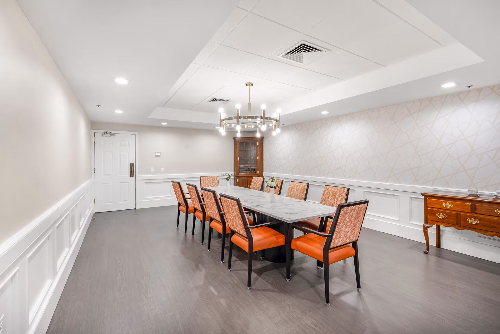 A bright, modern dining room with a long rectangular table surrounded by ten orange cushioned chairs. The room features white paneled walls, a patterned wallpaper accent wall, a wooden cabinet in the corner, a wooden sideboard with drawers, and a contemporary chandelier hanging from the ceiling.