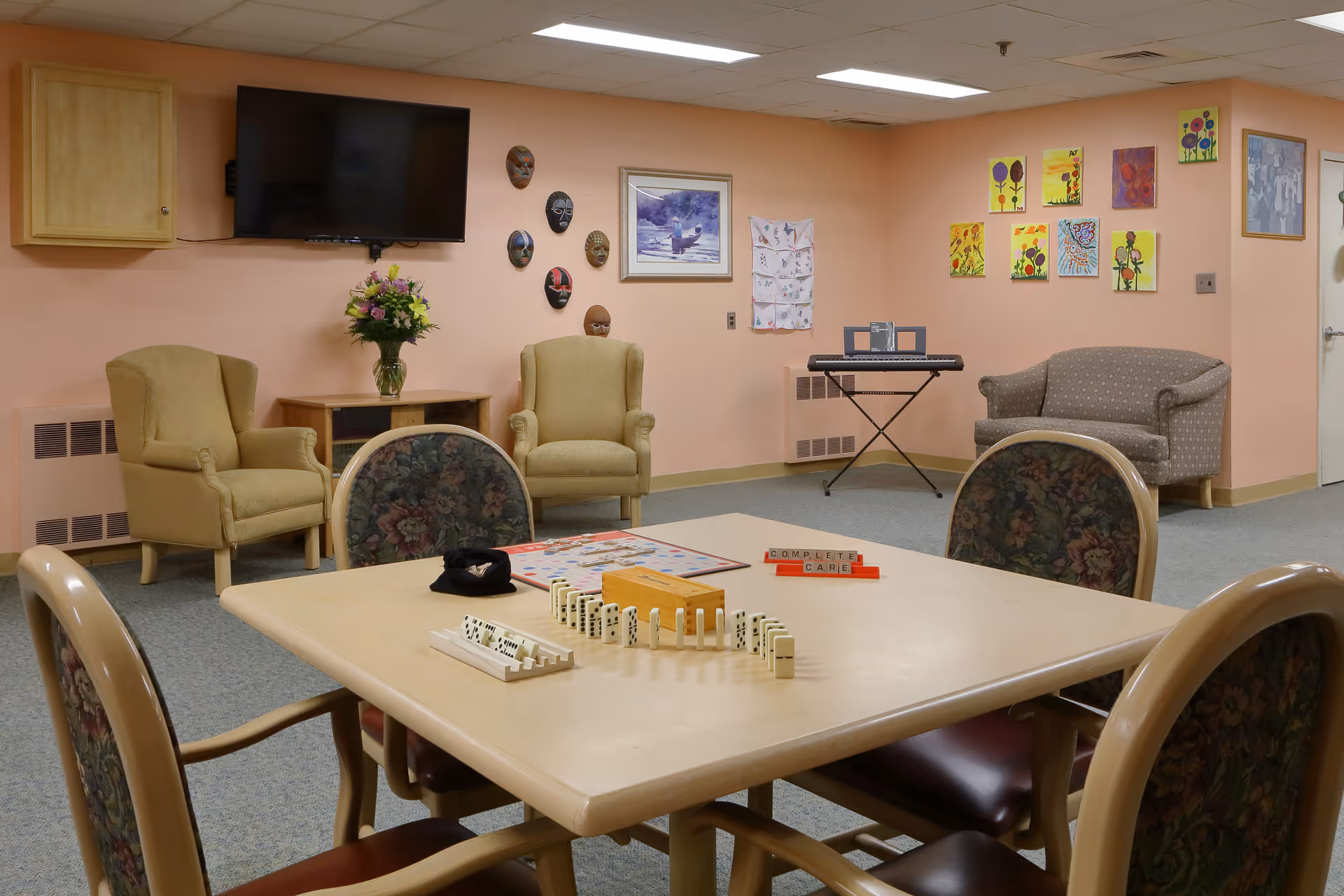 A cozy common room with a square table in the foreground set up for a game of dominoes and Scrabble. The room has floral upholstered chairs around the table, two beige armchairs, a small couch, a keyboard on a stand, and various colorful paintings and masks on peach-colored walls. A flat-screen TV is mounted on the wall above a small cabinet with a vase of flowers.