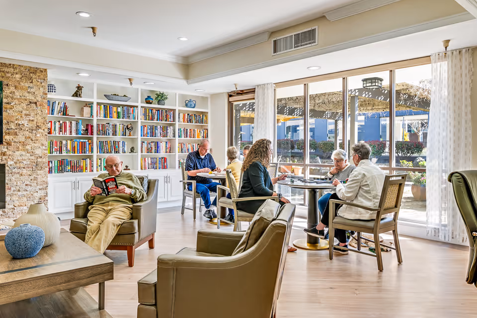 A bright and spacious common area in a retirement center with large windows letting in natural light. Several elderly people are seated, some reading books and others engaged in conversation around a table. The room features a built-in bookshelf filled with books, comfortable armchairs, and light wood flooring.
