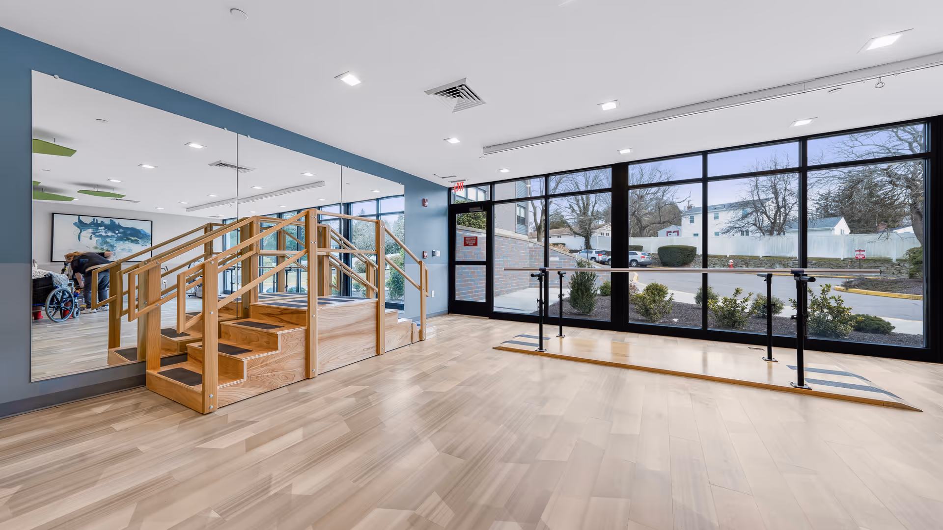 A rehabilitation therapy room with wooden stairs and handrails for physical therapy exercises. The room has large floor-to-ceiling windows letting in natural light, a mirrored wall on one side, and light wood flooring. Outside, there are shrubs and a parking area visible through the windows.