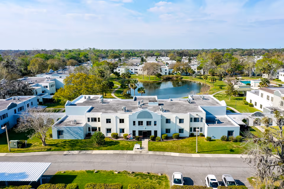 Aerial view of white two-story senior living buildings facing a pond with landscaped grounds and parking.
