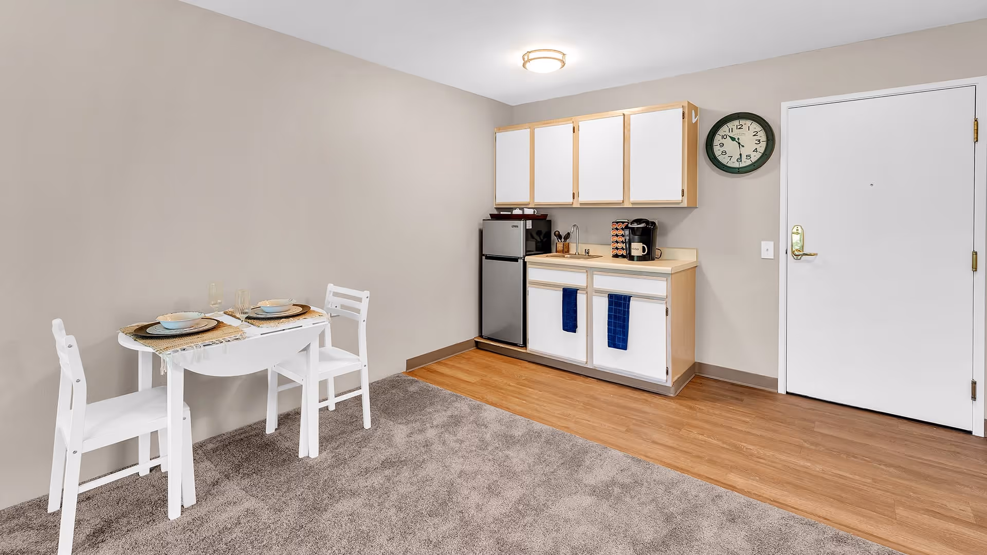 Small kitchenette area with white cabinets, a mini refrigerator, a coffee maker, and a sink. Next to the kitchenette is a small white dining table set for two with plates, bowls, and glasses. The floor is a combination of carpet and wood, and there is a wall clock above the kitchenette near a white door.