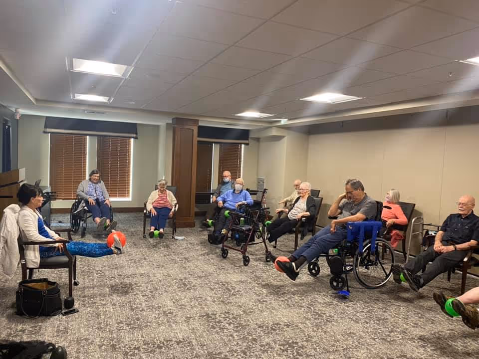 A group of elderly people seated in a circle in a carpeted room, participating in a seated exercise activity by lifting their legs. Some individuals are using wheelchairs or walkers. The room has beige walls, a drop ceiling with fluorescent lights, and windows with closed blinds.