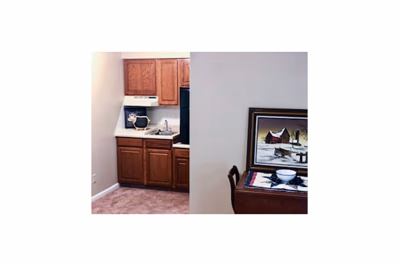 Small kitchen area with wooden cabinets, a sink, a stove with a range hood, and a black refrigerator. Adjacent to the kitchen is a wooden table with a chair, a bowl on a colorful table runner, and a framed painting of a snowy rural scene on the wall.