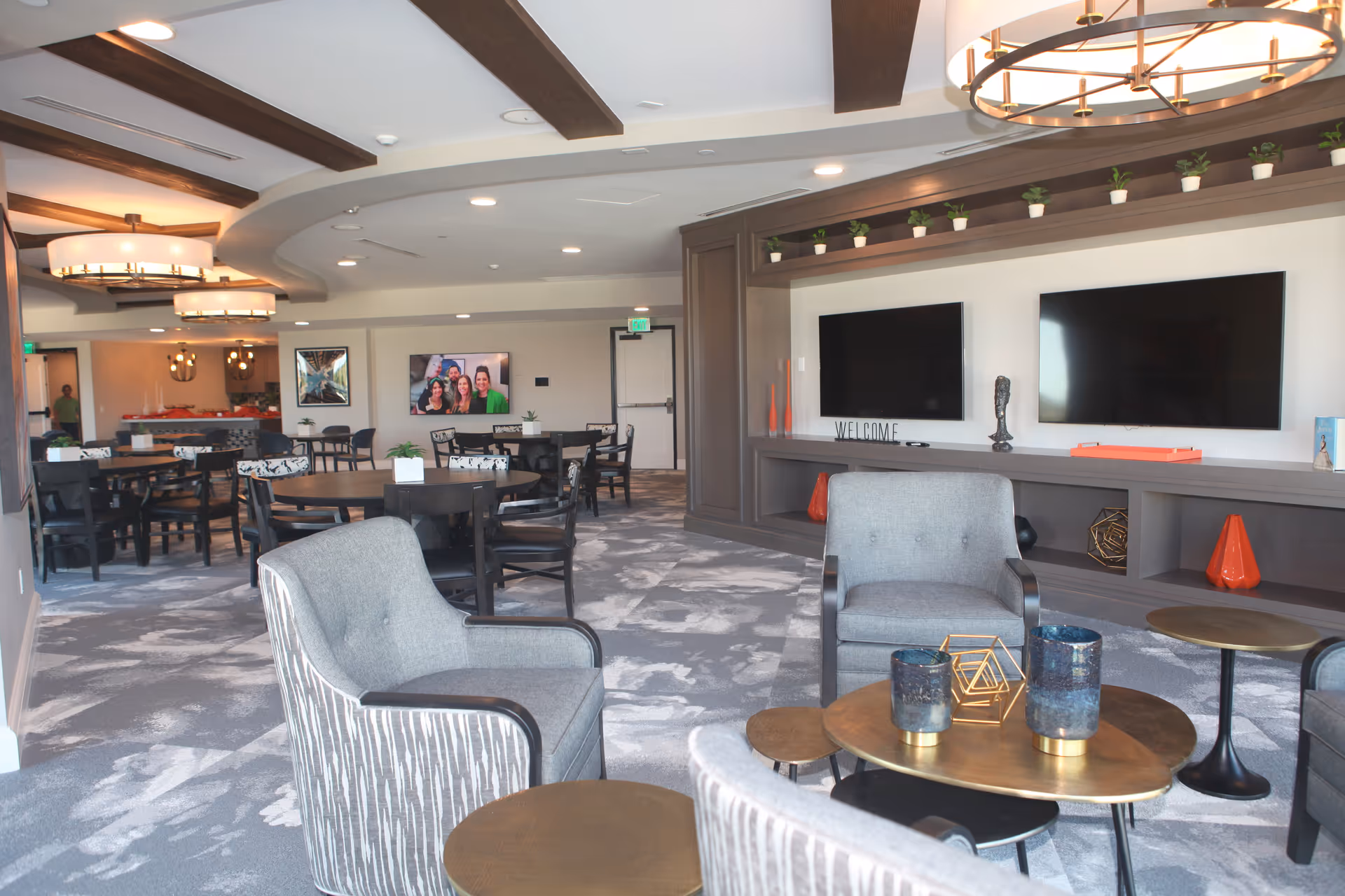 A spacious and modern common area with gray armchairs and round wooden tables in the foreground. The room features a patterned carpet, multiple round dining tables with chairs, two large flat-screen TVs mounted on a wall with decorative shelves above, and contemporary ceiling lights with wooden beams. There are small potted plants on the shelves and tables, and a welcoming atmosphere is created by the decor and lighting.