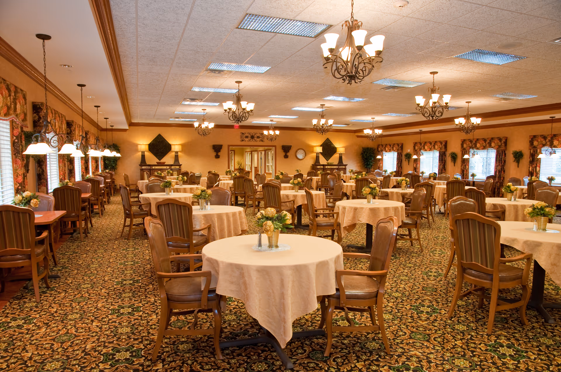 A spacious dining room with multiple round tables covered with beige tablecloths, each adorned with a small floral centerpiece. The room features wooden chairs with armrests, patterned carpet, and several chandeliers hanging from the ceiling. Large windows with floral curtains line the walls, allowing natural light to enter. The overall decor is warm and inviting.