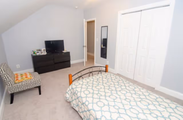 A bedroom with a single bed covered with a patterned blue and white bedspread. There is a black dresser with a small TV on top, a patterned armchair with a colorful cushion, a wall-mounted mirror, and a closet with white double doors. The walls are painted light gray and the carpet is beige.