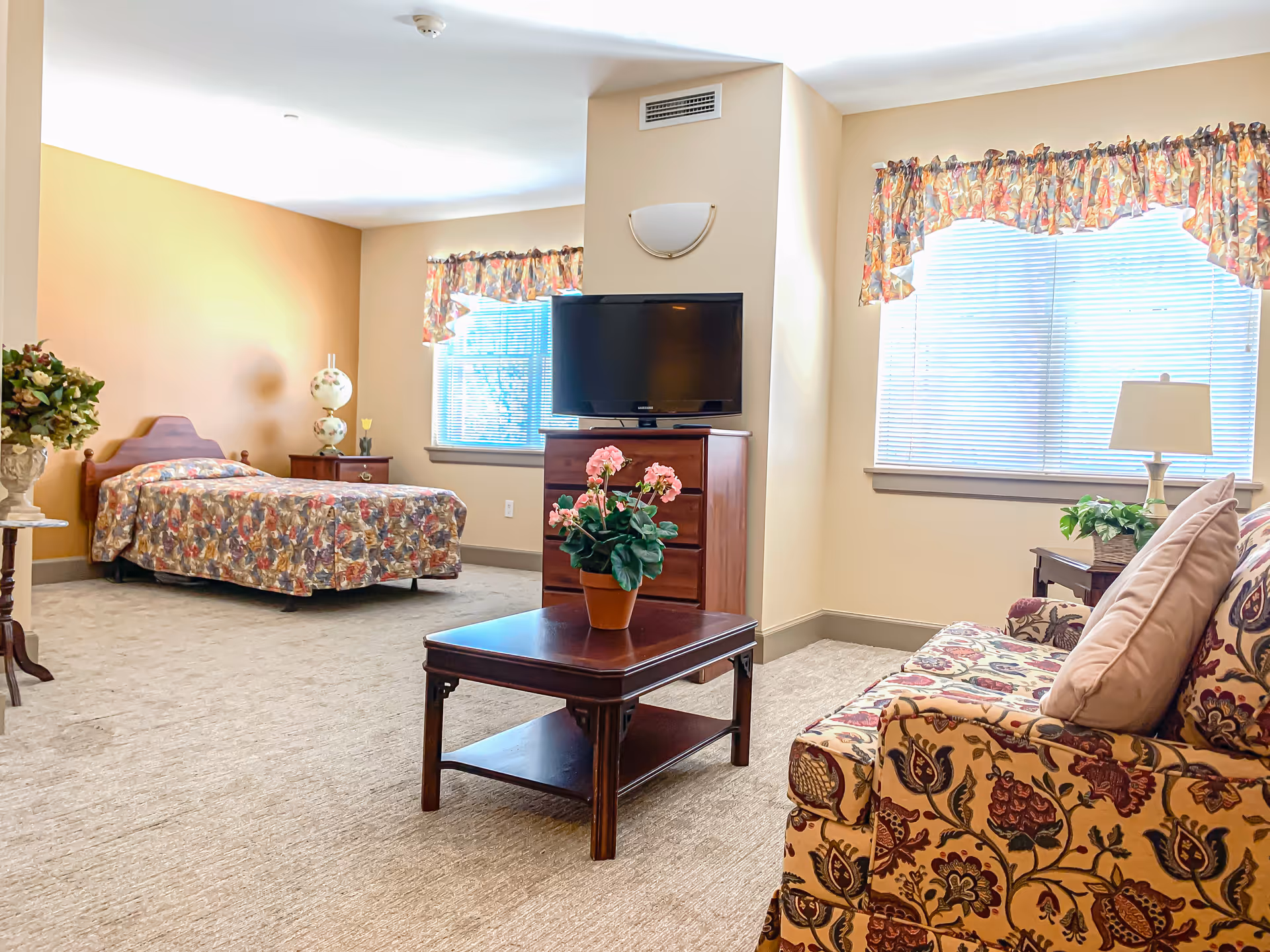 A bright and spacious senior living room with a single bed covered in a floral bedspread, a wooden nightstand with a decorative lamp, a wooden dresser with a flat-screen TV on top, a wooden coffee table with a potted pink flower, and a floral patterned sofa with cushions. The room has two windows with blinds and floral valances, beige walls, and carpeted floor.