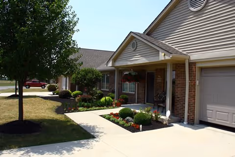 Exterior view of a single-story residential building with beige siding and brick accents. The entrance features a covered porch with a bench and hanging flower baskets. There is a well-maintained garden with shrubs and flowers along the walkway, and a tree on the lawn beside the sidewalk. A red vehicle is parked in the background under a clear blue sky.