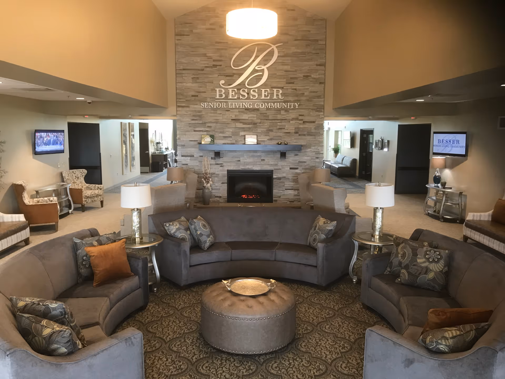 Lobby lounge of a senior living community with curved sofas around a central ottoman, a fireplace and a stone feature wall bearing the Besser Senior Living Community name.