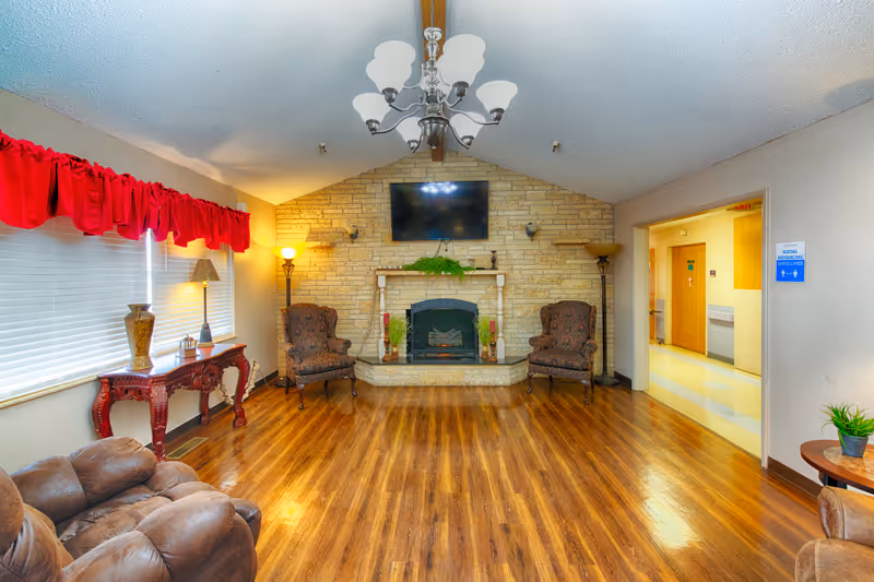 Common area living room with hardwood floors, a stone fireplace topped by a TV, upholstered chairs, and a chandelier.