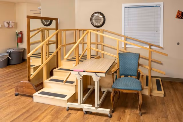 A small wooden staircase with handrails on both sides, designed for physical therapy or rehabilitation, with a green upholstered chair and a mobile table in front. There is a large mirror on the left wall, a clock on the wall above the stairs, a window with closed blinds on the right, and a fire extinguisher and trash bins in the background.