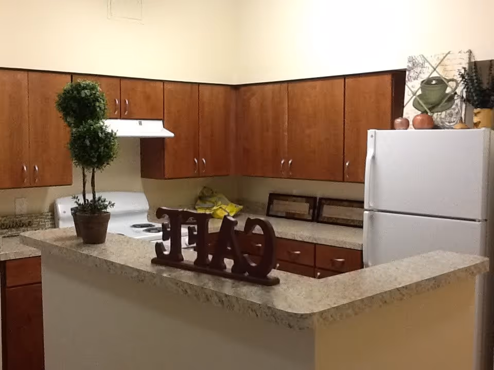 Interior view of a kitchen with wooden cabinets, a white stove with a range hood, a white refrigerator, and a countertop with a decorative sign spelling 'EAT'. There is a potted plant on the counter and some framed pictures and decorative items on the countertop near the refrigerator.