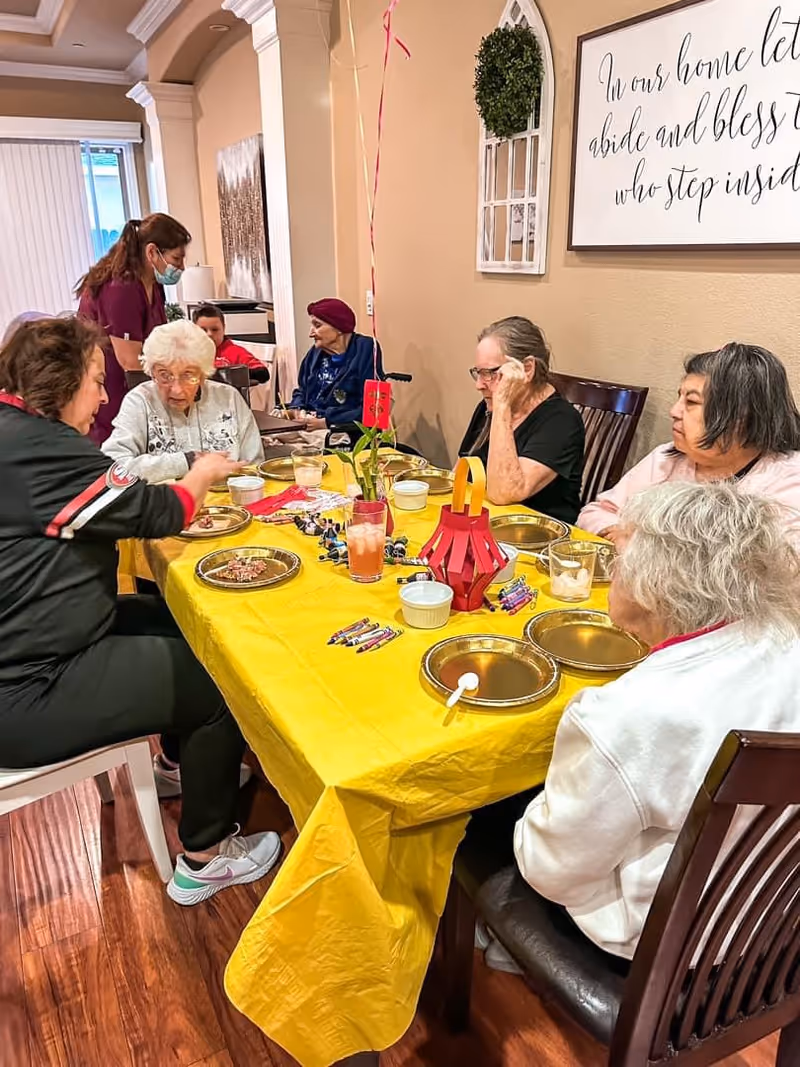 A group of elderly women sitting around a dining table covered with a yellow tablecloth, engaging in conversation and eating. A caregiver wearing a mask is attending to one of the women. The room has beige walls with decorative wall art and a small plant centerpiece on the table.