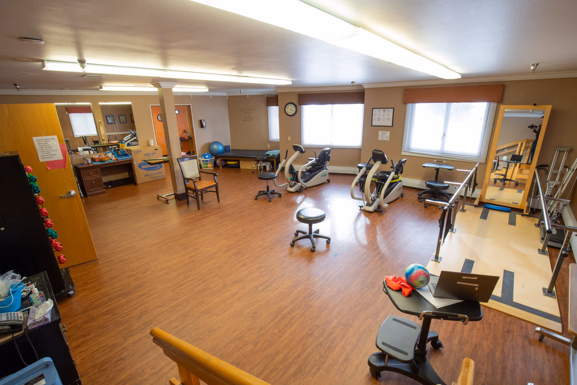 A spacious rehabilitation room with exercise equipment including stationary bikes, parallel bars for walking practice, chairs, a large mirror, and various therapy tools. The room has wood flooring, beige walls, and large windows letting in natural light.