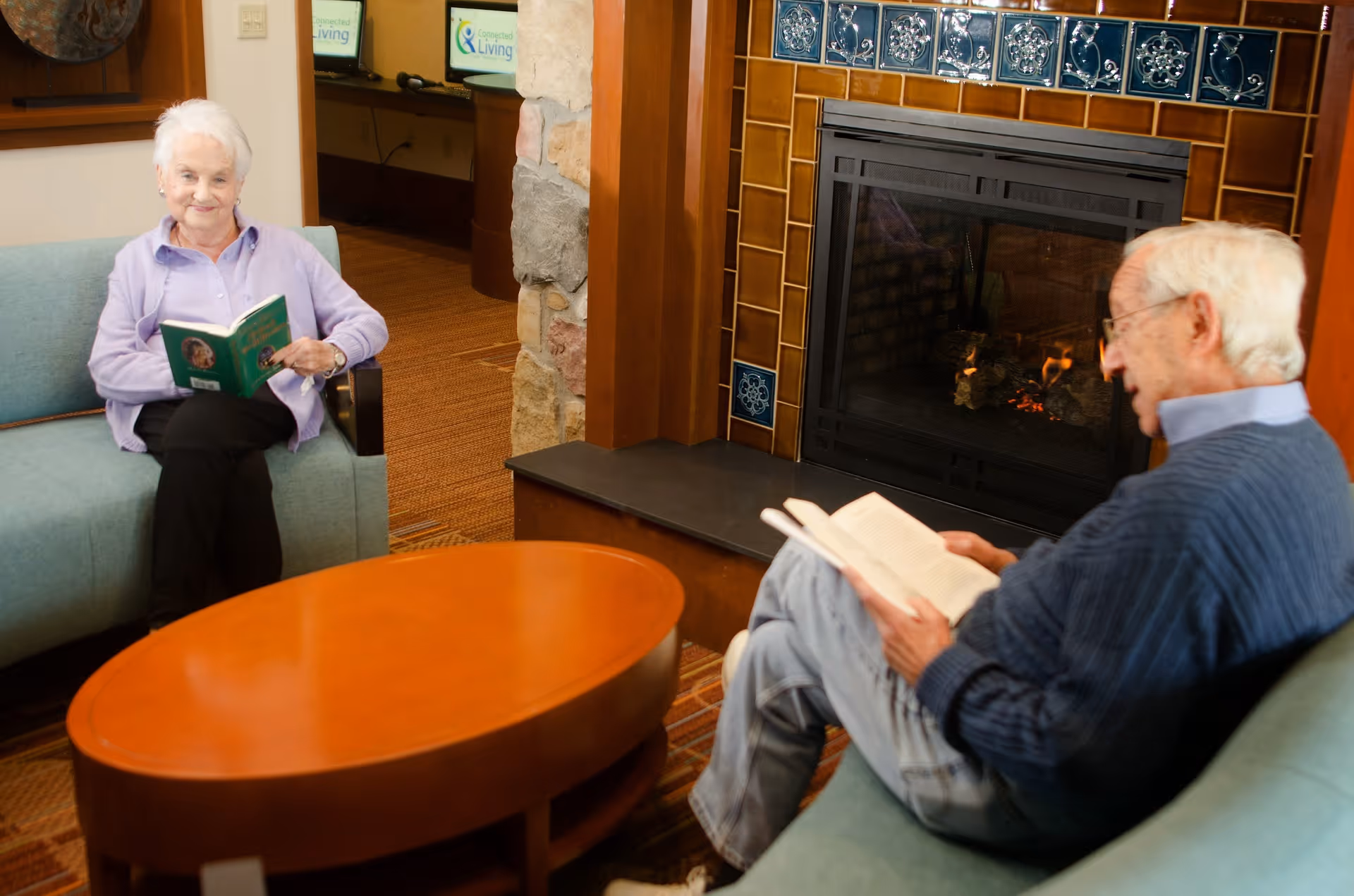 An elderly woman and an elderly man sitting on separate couches in a cozy living room area, both reading books. There is a wooden coffee table between them and a fireplace with decorative tiles in the background.