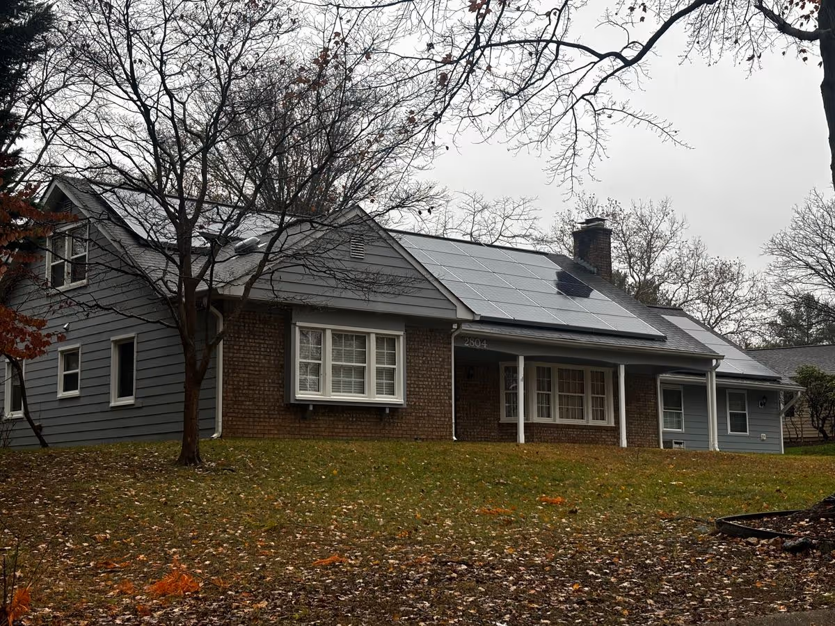 Brick and gray-sided single-story house with solar panels on the roof and a grassy front yard with bare trees.