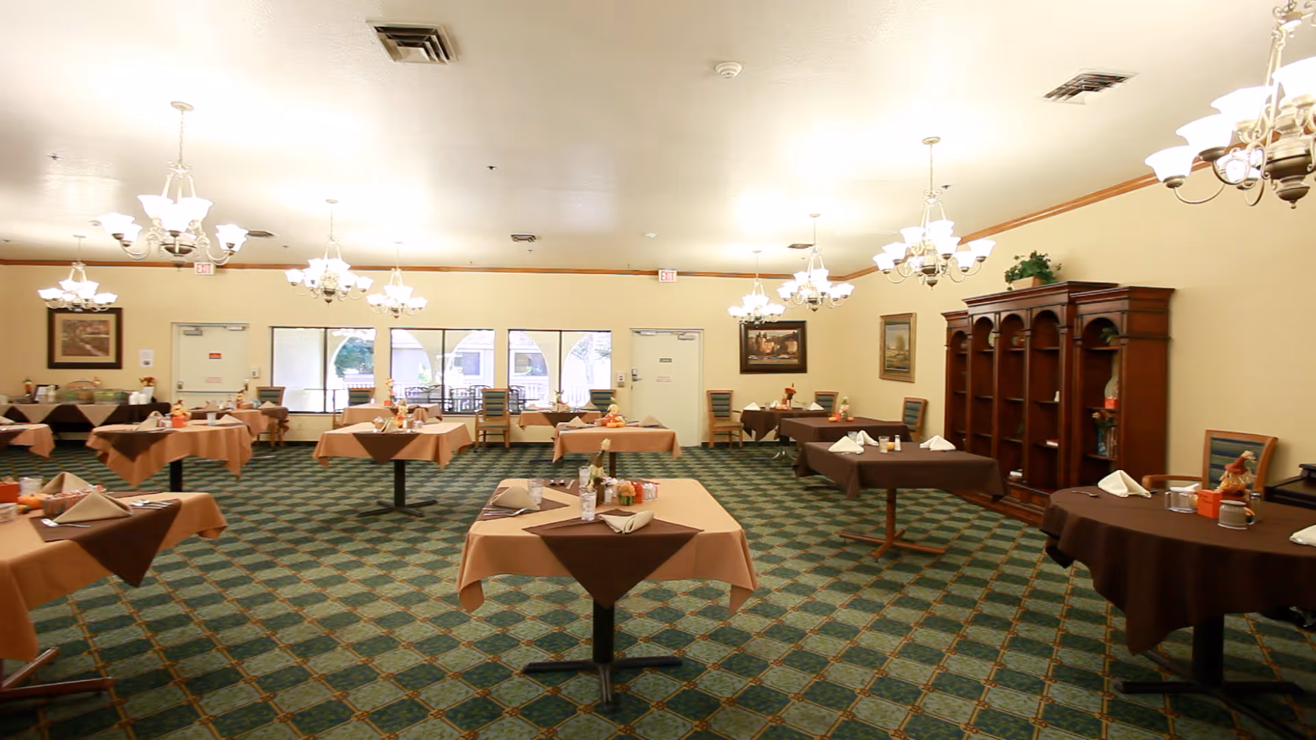 A spacious dining room with multiple tables covered in brown and beige tablecloths, each set with napkins, condiments, and small decorations. The room has green patterned carpet, cream-colored walls, several chandeliers hanging from the ceiling, and large windows letting in natural light. There is a wooden cabinet along one wall and framed artwork on the walls.