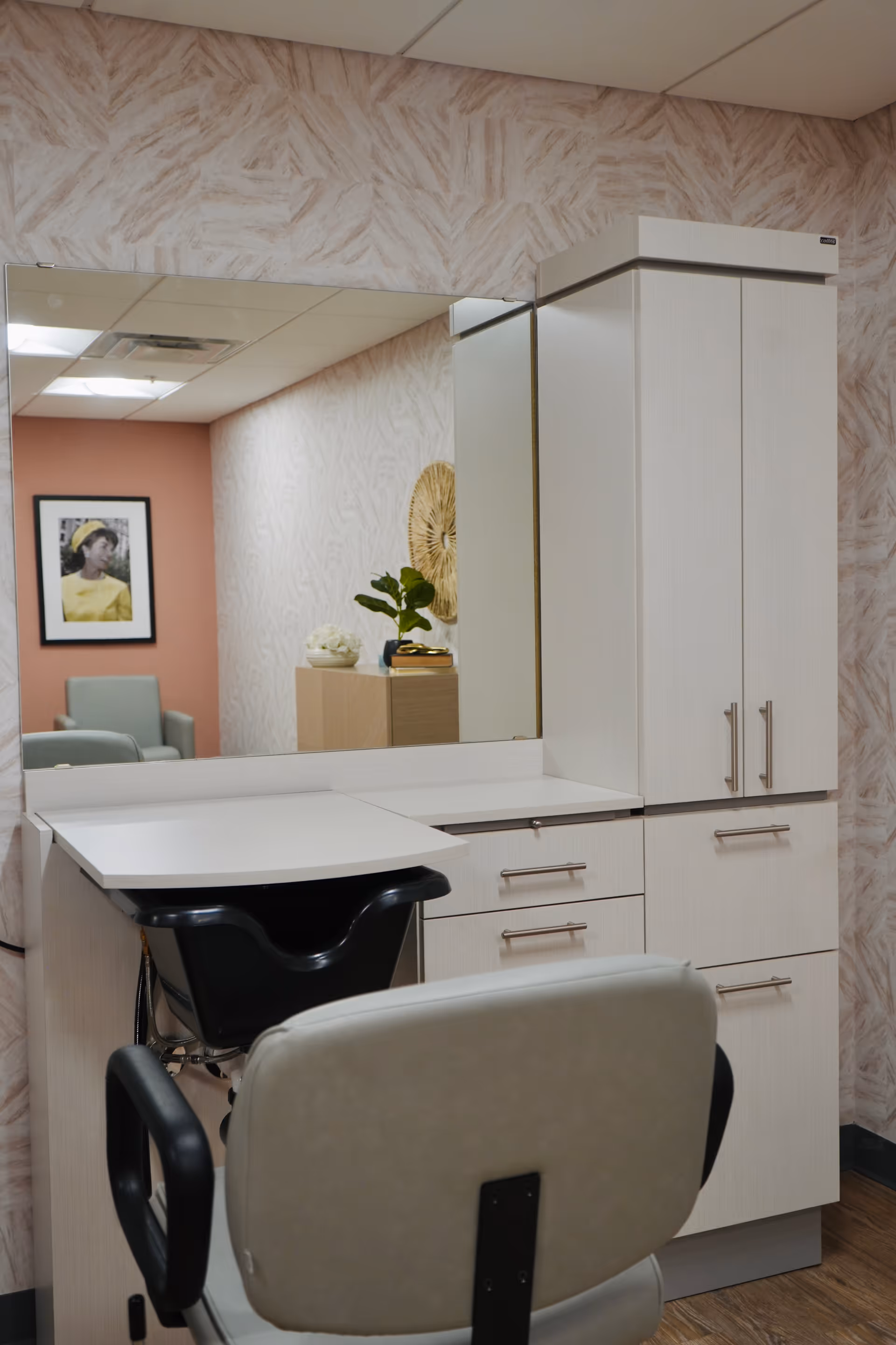 Hair-washing salon station with a padded chair, shampoo basin, large mirror, and white storage cabinets.