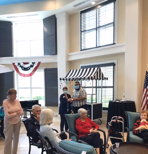 A group of elderly people sitting and standing in a bright, spacious common area with large windows and high ceilings. Two staff members wearing masks stand near a small cart with a striped canopy. The room is decorated with patriotic bunting and an American flag is visible in the corner.