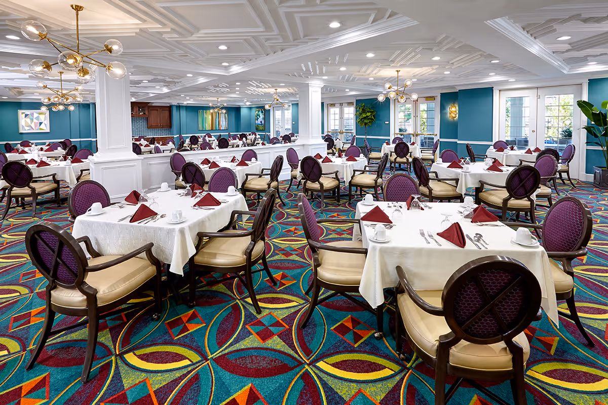 A spacious dining room with multiple tables covered in white tablecloths, each set with maroon folded napkins, cups, and silverware. The chairs have dark wooden frames with purple cushioned backs and beige seats. The room features a colorful geometric carpet, teal walls, white ceiling with recessed lighting, and modern gold chandeliers. Large windows allow natural light to enter the space.
