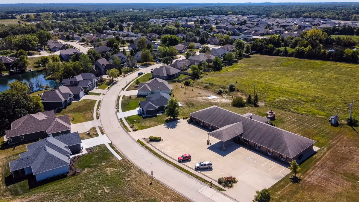 Aerial view of a single-story senior living building with a parking lot, surrounded by suburban houses and open fields.