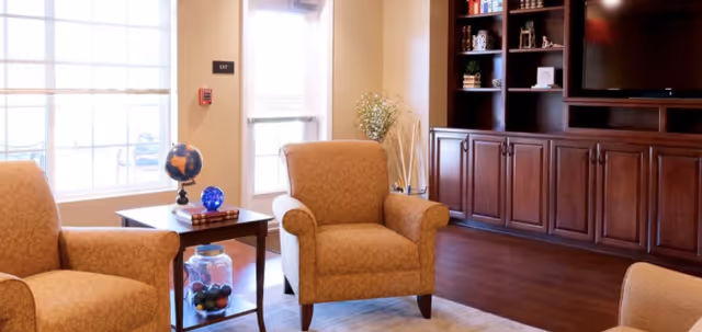 Sunny common living area with upholstered armchairs, a side table holding a globe and decorative items, and built-in wooden cabinets with a TV.