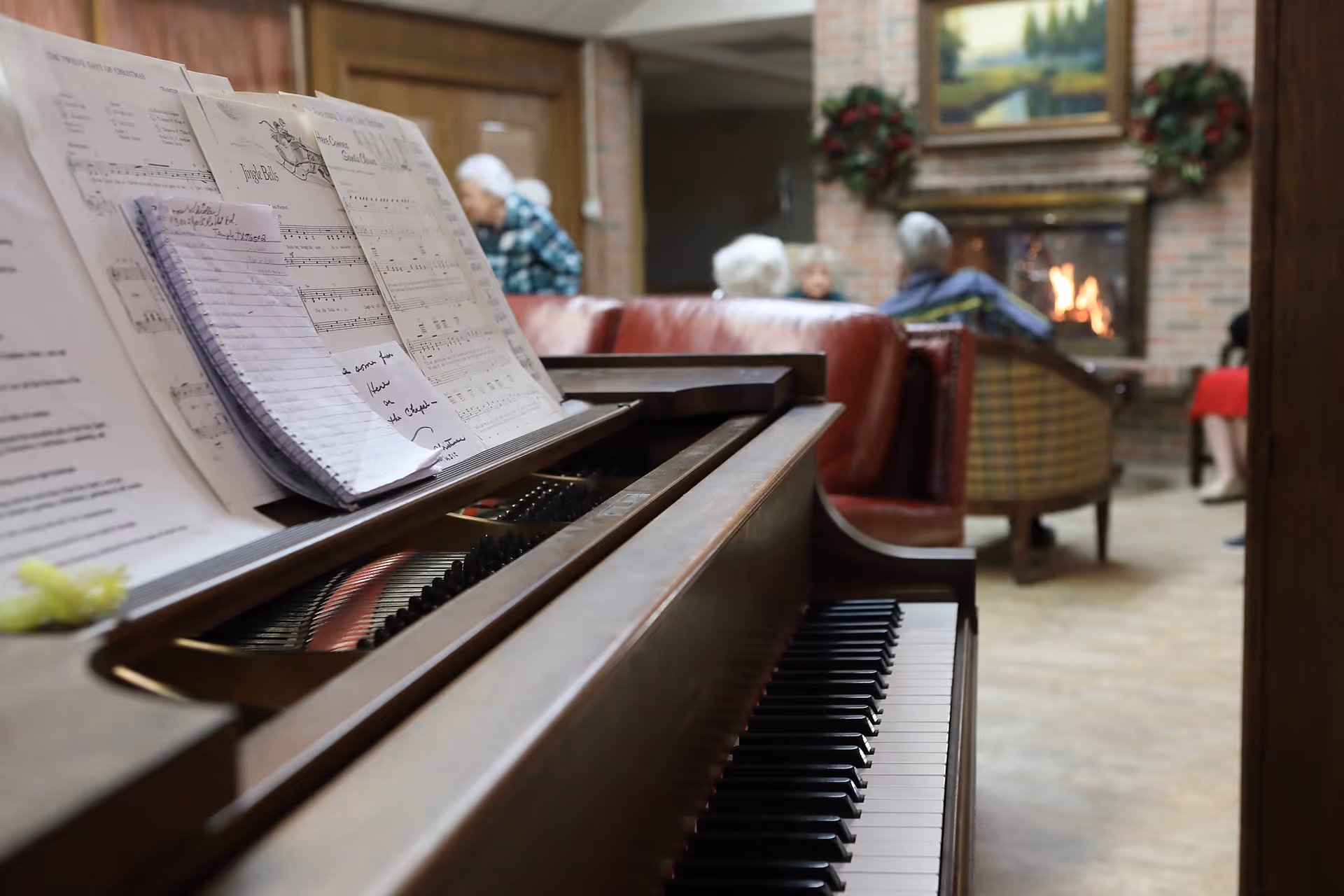 Close-up view of a piano keyboard with sheet music on the stand in a cozy room. In the background, several elderly people are seated on sofas and chairs near a lit fireplace with a painting and wreaths on the brick wall.