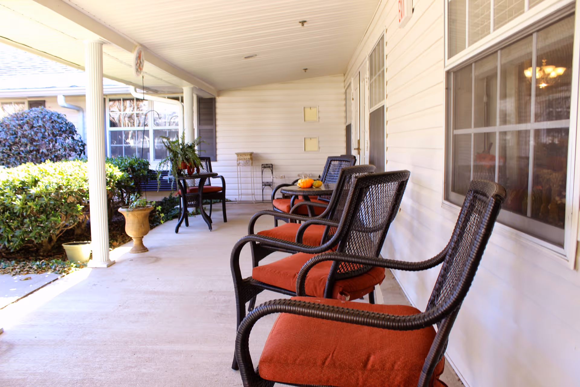 Covered outdoor porch area with several black wicker chairs with red cushions arranged along the wall of a white building. There are small tables with plants and decorative items, and bushes and greenery are visible outside the porch.