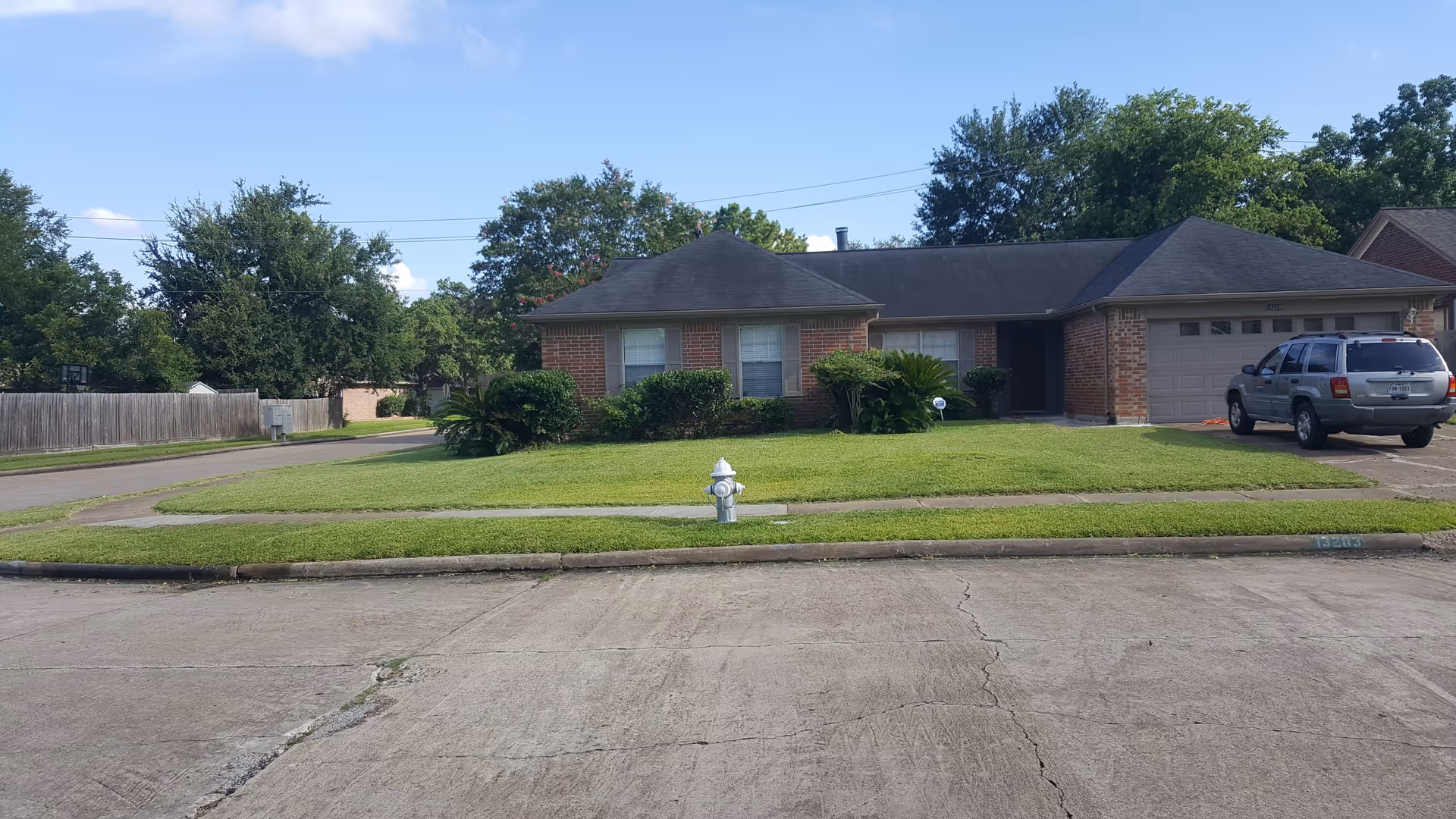 Front exterior of a one-story brick suburban house with a lawn, driveway, a parked SUV, and a fire hydrant in the foreground.