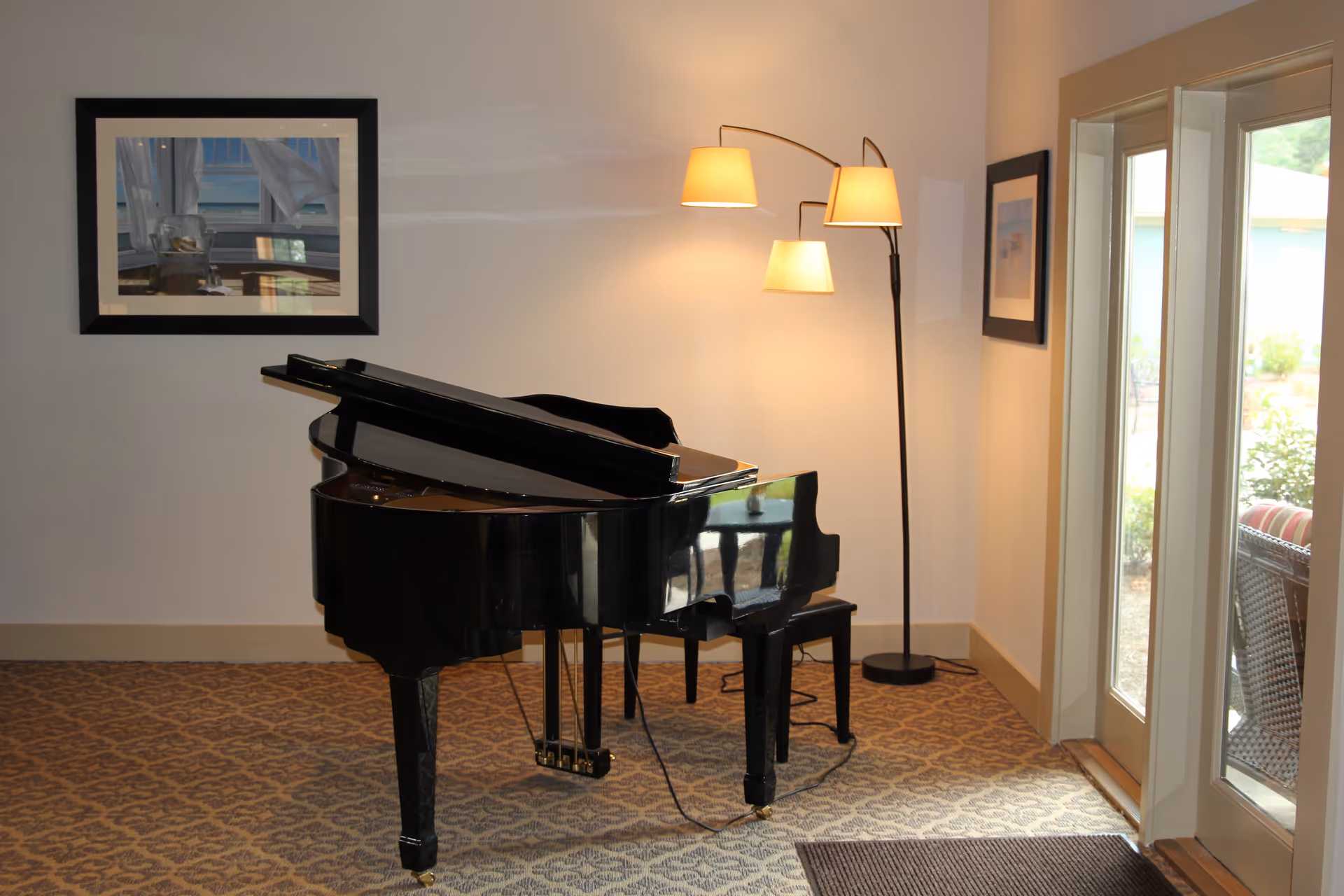 Interior room with a black grand piano and matching bench on a patterned carpet. A three-lamp floor light is turned on near the piano. Two framed pictures hang on the walls, and there are glass doors leading outside where some outdoor furniture is visible.