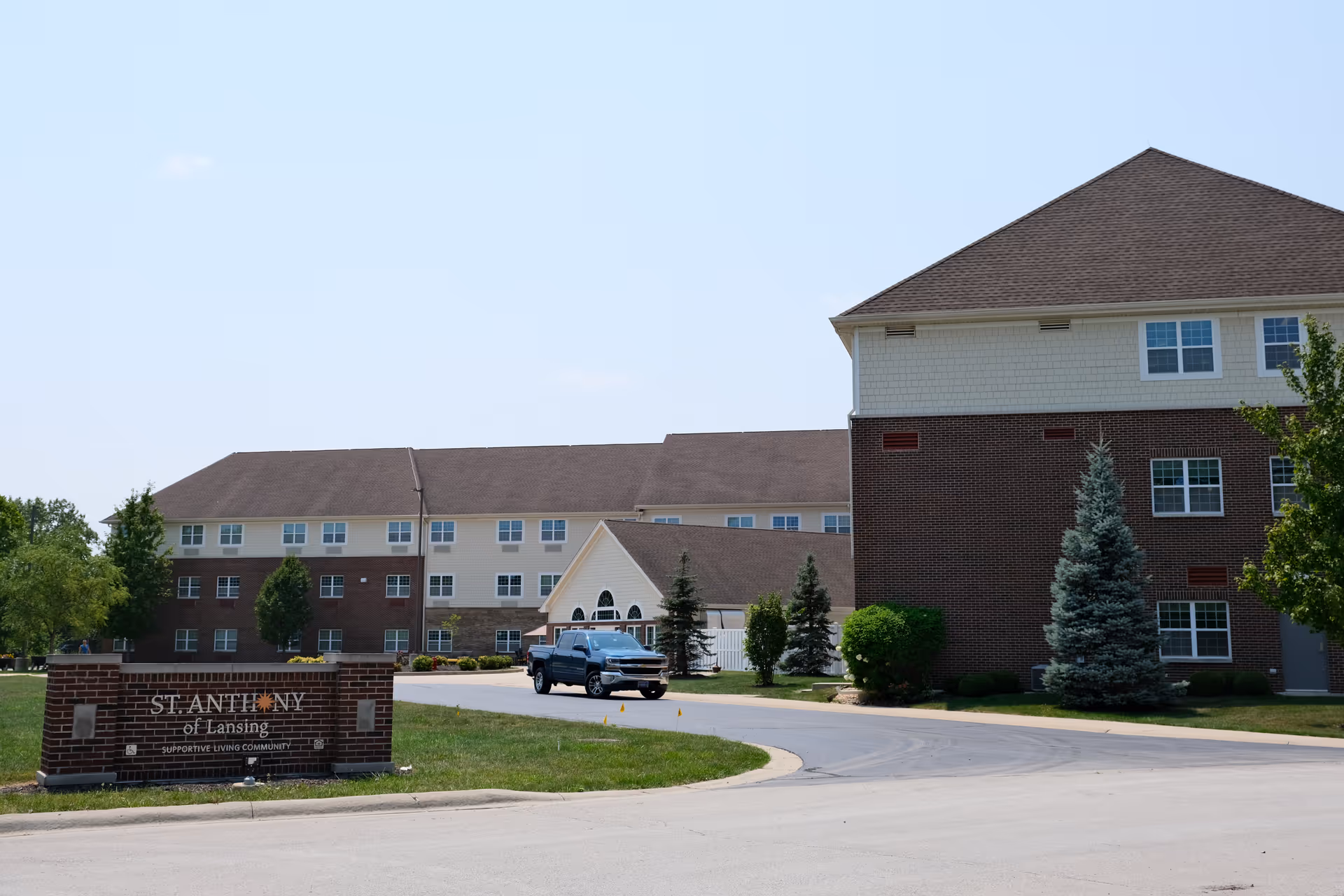 Front exterior of the St. Anthony of Lansing senior living building with a sign, driveway, and a parked truck.