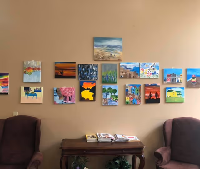 Two armchairs flank a wooden table with magazines beneath a wall display of small colorful paintings.