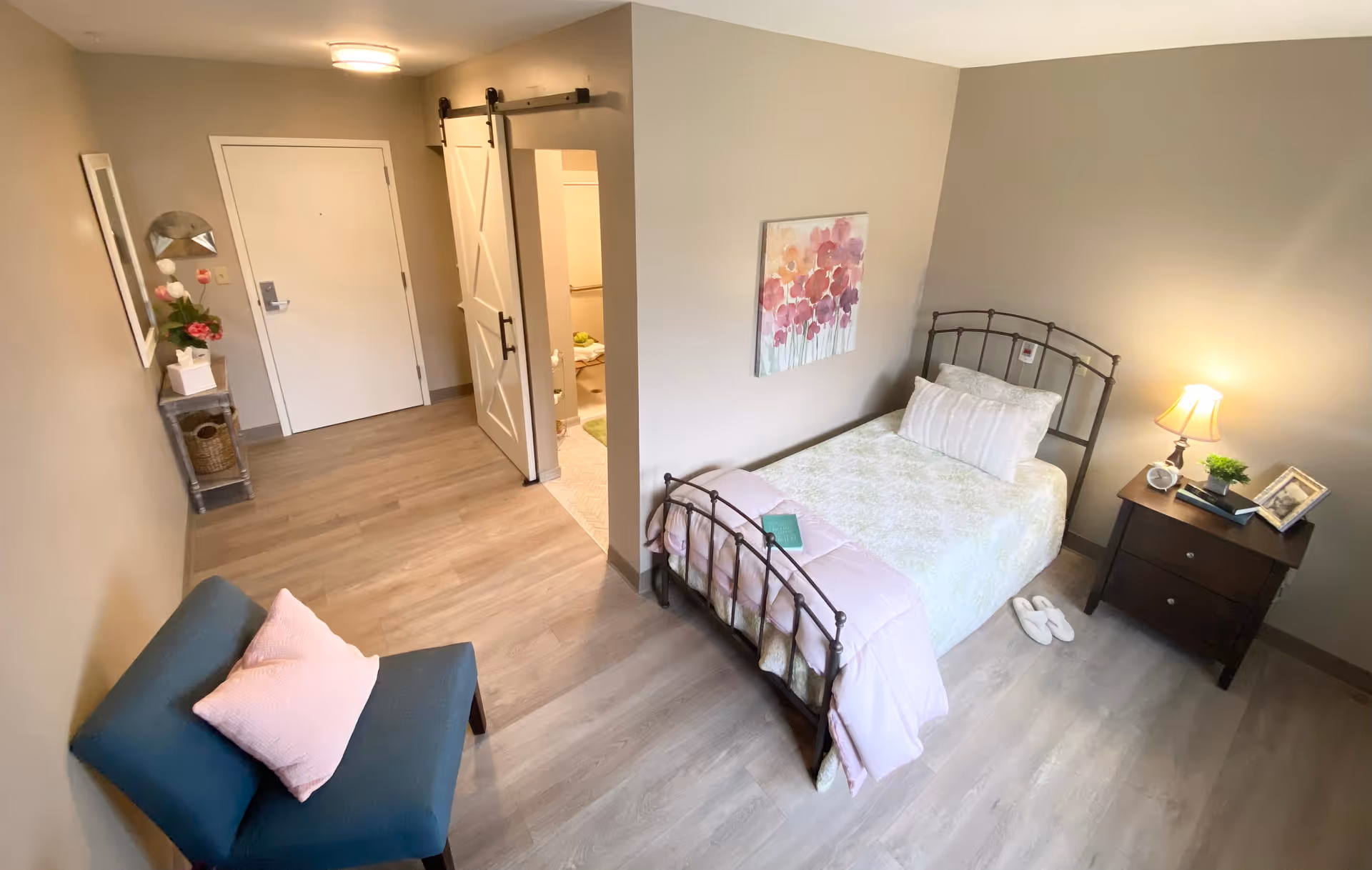 A cozy bedroom in a senior living facility featuring a single bed with light bedding and a pink throw, a dark wooden nightstand with a lamp, books, and a framed photo, a blue chair with a pink pillow, a small table with flowers and a mirror near the entrance door, and a sliding barn door leading to a bathroom.