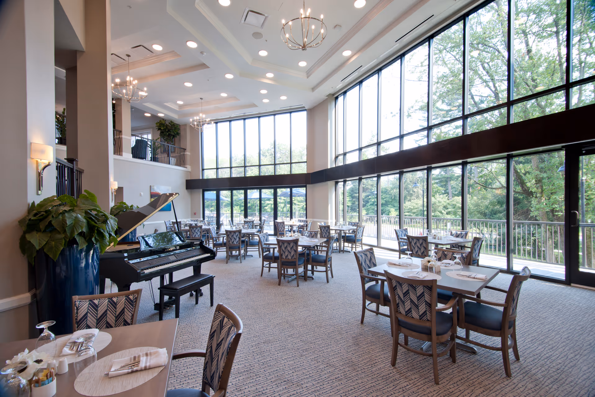 Bright double-height dining room with floor-to-ceiling windows, multiple set tables, and a grand piano near a large plant.